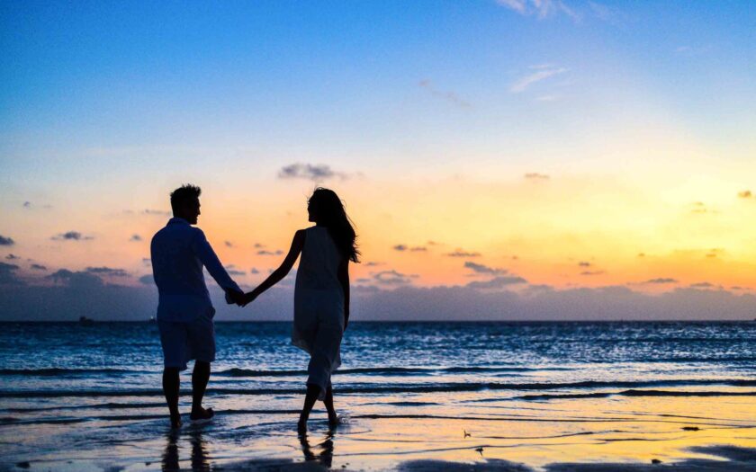 man and woman holding hands walking on seashore during sunrise